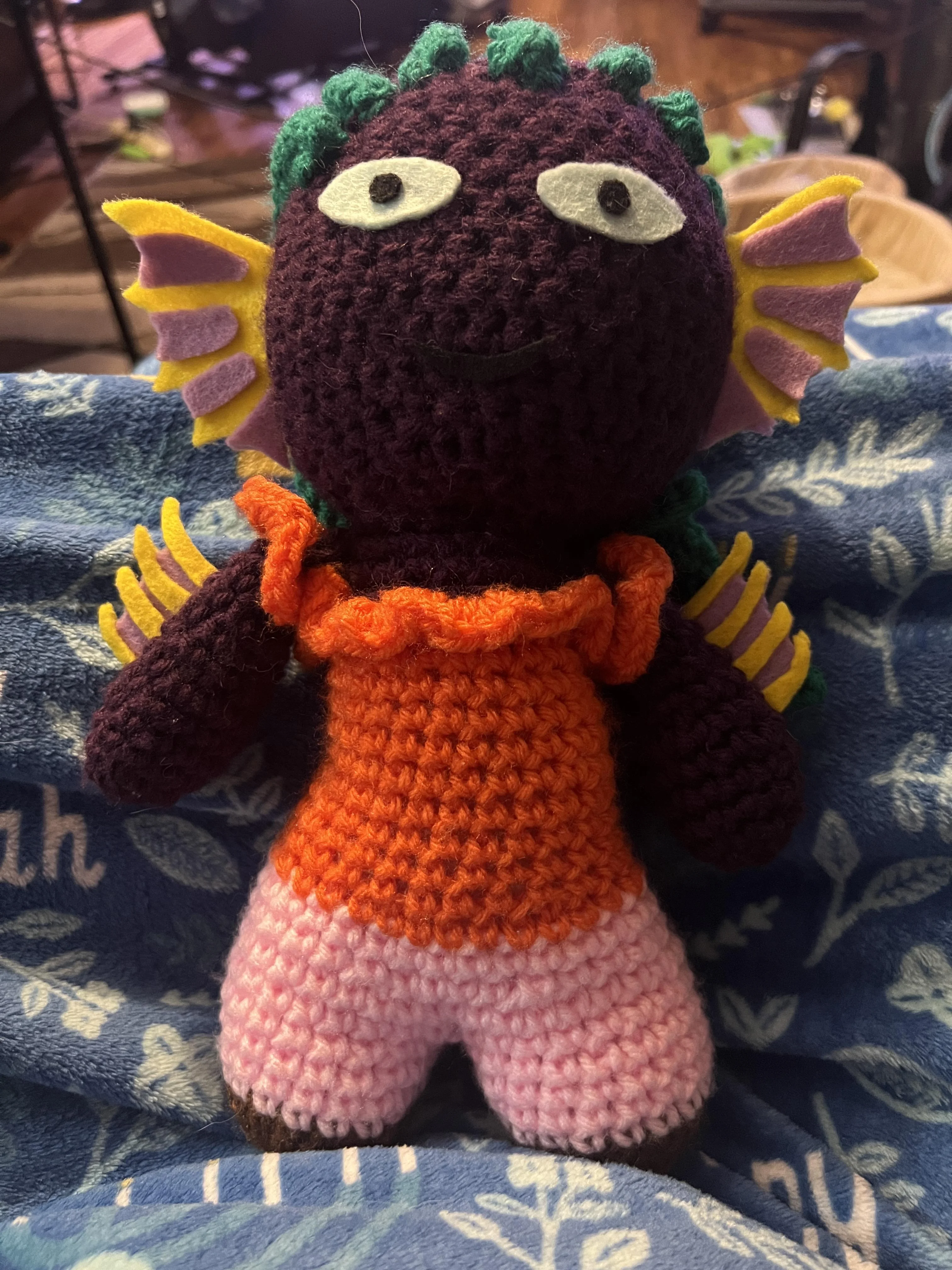 Purple skinned crochet doll with green braids and orange ruffly shirt